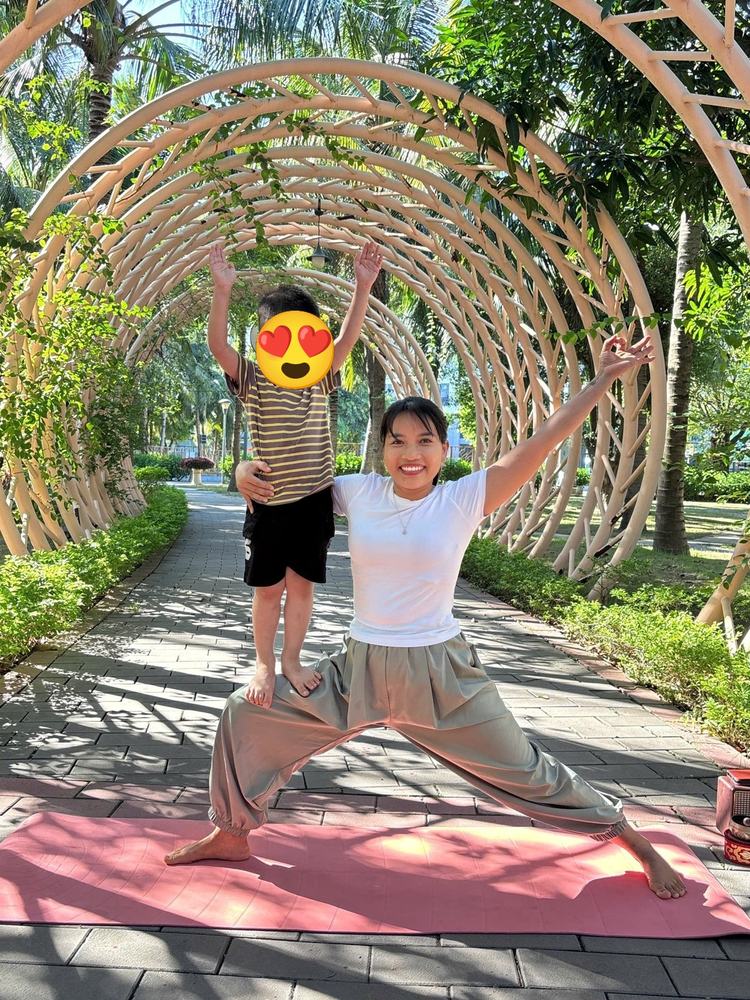 Cô Sophia tư thế chiến binh, bé trai đứng trên đùi cô giơ cao hai tay trong đường hầm cây xanh Ecopark